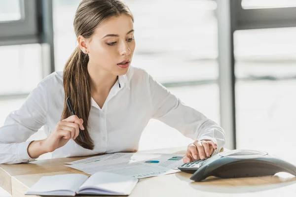 Jeune femme d'affaires confiante avec la paperasserie en utilisant le téléphone de conférence au bureau — Photo de stock