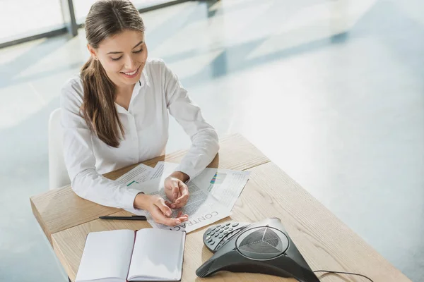 Blick aus der Vogelperspektive auf junge, glückliche Geschäftsfrau, die Papierkram erledigt und per Konferenztelefon auf dem Tisch im Büro spricht — Stockfoto