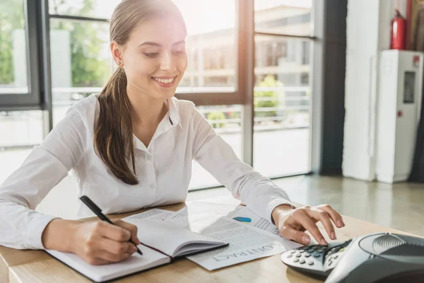 Junge lächelnde Geschäftsfrau erledigt Papierkram und drückt Taste des Konferenztelefons auf dem Tisch im Büro — Stockfoto