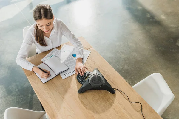 Hochwinkelaufnahme einer jungen, glücklichen Geschäftsfrau, die Papierkram erledigt und den Knopf ihres Konferenztelefons auf dem Tisch im Büro drückt — Stockfoto