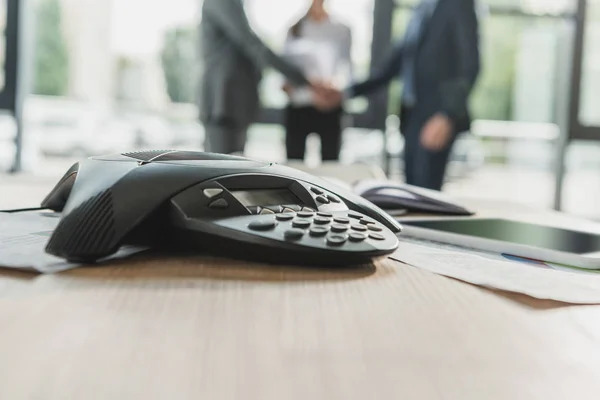 Close-up shot of conference phone with blurred business people shaking hands on background at modern office — Stock Photo