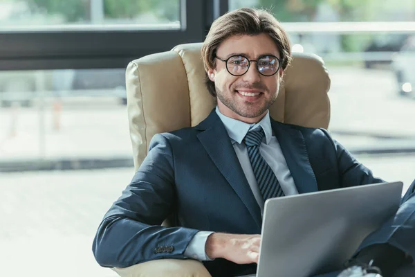 Guapo sonriente hombre de negocios sentado usando el ordenador portátil en sillón y mirando a la cámara - foto de stock