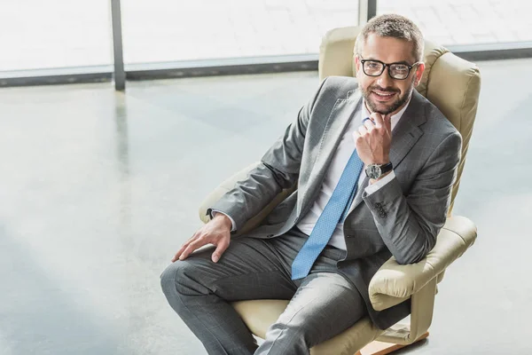 Visão de alto ângulo de belo homem de negócios sorridente sentado em poltrona de luxo no escritório moderno — Fotografia de Stock