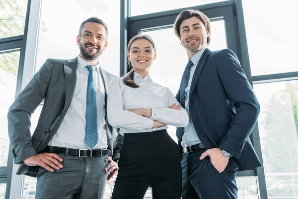 Вид снизу группы улыбающихся молодых бизнесменов, стоящих в современном офисе и смотрящих в камеру — стоковое фото