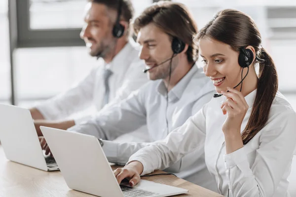 Sorrindo gerentes de call center em camisas brancas trabalhando juntos enquanto sentados em fila no escritório moderno — Fotografia de Stock