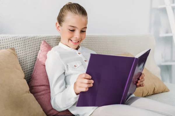 Entzückendes kleines Kind liest Buch, während es sich zu Hause auf der Couch entspannt — Stockfoto