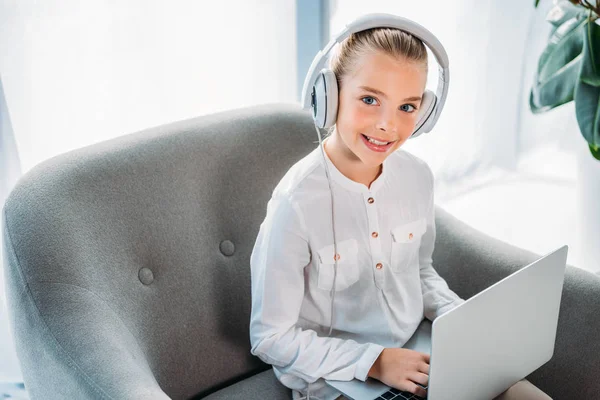 Lächelndes kleines Kind mit Kopfhörer und Laptop, während es im Sessel sitzt und in die Kamera schaut — Stockfoto
