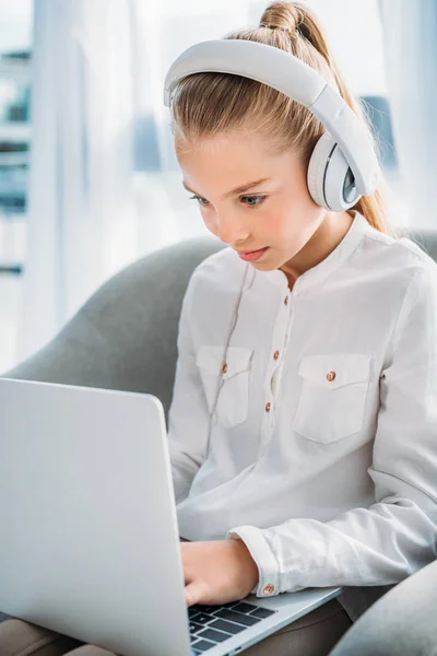Concentrated little child in headphones using laptop while sitting in armchair — Stock Photo