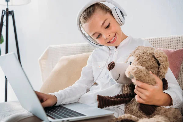 Adorable petit enfant dans les écouteurs en utilisant un ordinateur portable tout en étant assis dans un fauteuil avec ours en peluche — Photo de stock