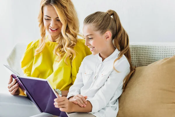 Feliz madre e hija sentadas en el sofá y leyendo el libro en casa - foto de stock