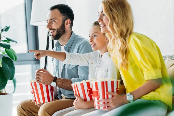 Feliz joven familia viendo la película en casa con cubos de palomitas de maíz y apuntando a la televisión - foto de stock