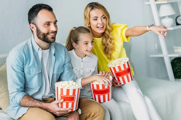 Sorrindo jovem família assistindo filme em casa com baldes de pipoca e apontando para a tv — Fotografia de Stock