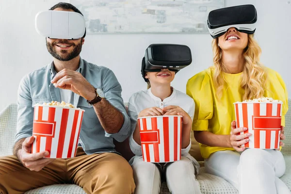 Happy young family in virtual reality headsets watching movie with buckets of popcorn — Stock Photo