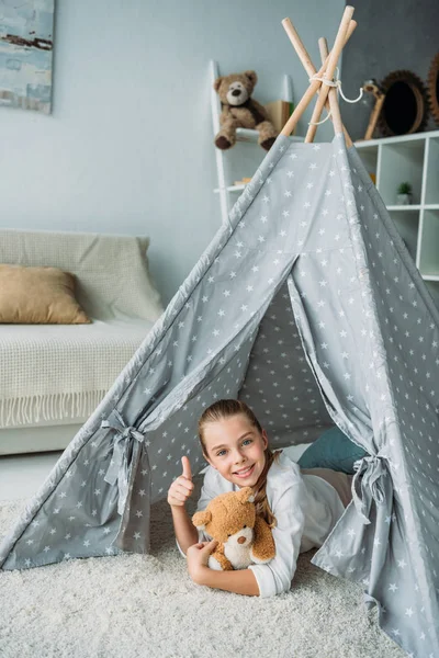 Adorable little child lying on floor in teepee with teddy bear and showing thumb up at home — Stock Photo