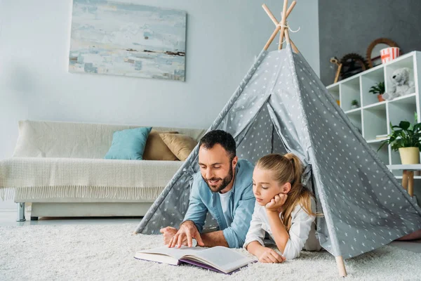 Father and daughter lying on floor inside of teepee and reading book together at home — Stock Photo