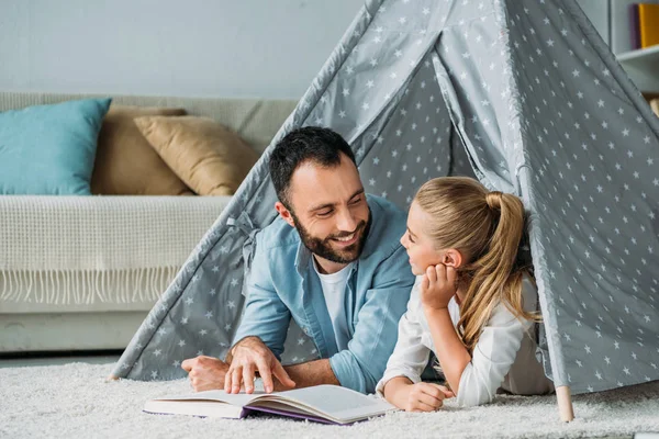 Happy father and daughter lying on floor inside of teepee and reading book together at home — Stock Photo