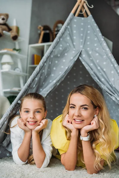 Madre e hija tumbadas en tipi y mirando a la cámara en casa - foto de stock