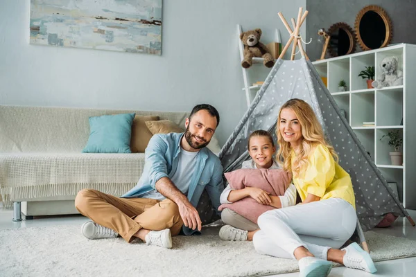 Happy young family sitting on floor with teepee and looking at camera — Stock Photo