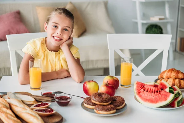 Adorable niño pequeño sentado a la mesa con varios alimentos para el desayuno en casa - foto de stock