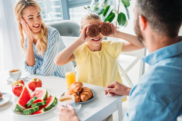 Sorrindo jovens pais olhando para sua filha cobrindo os olhos com donuts — Fotografia de Stock