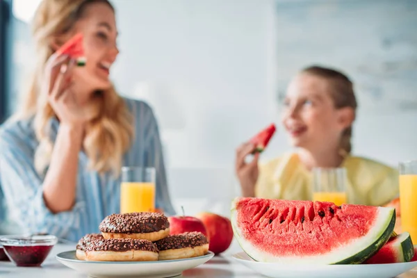 Foco seletivo tiro de mãe e filha tomando café da manhã com donuts e melancia em primeiro plano em casa — Fotografia de Stock