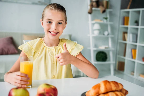 Lächelndes kleines Kind mit einem Glas Orangensaft, das den Daumen in die Kamera zeigt — Stockfoto