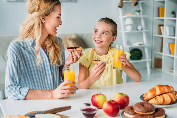 Mãe feliz e filha comendo baguete com geléia e suco de laranja em casa — Fotografia de Stock