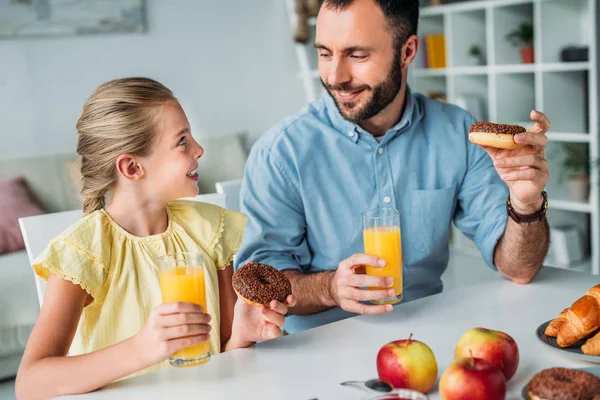 Feliz padre e hija comiendo donas con jugo de naranja en casa - foto de stock