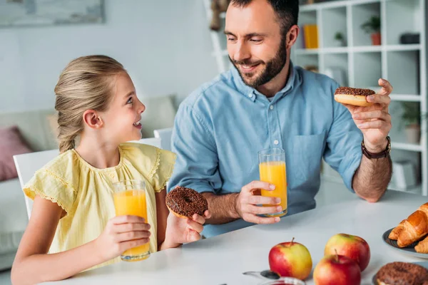 Pai feliz e filha comendo donuts com suco de laranja em casa — Fotografia de Stock