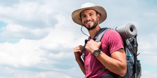 Smiling traveler in hat with backpack and tourist mat, with cloudy sky background — Stock Photo