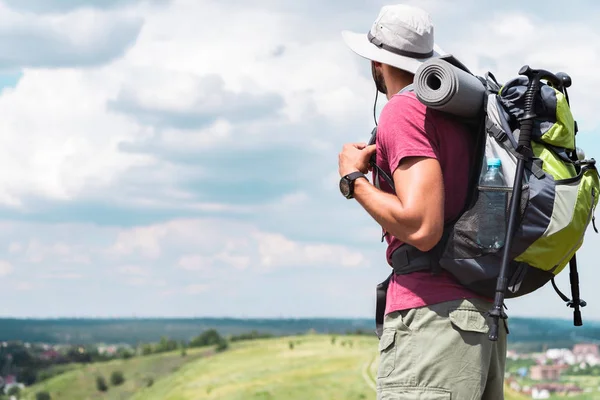 Viajero masculino en sombrero con mochila y alfombra turística mirando el prado de verano - foto de stock