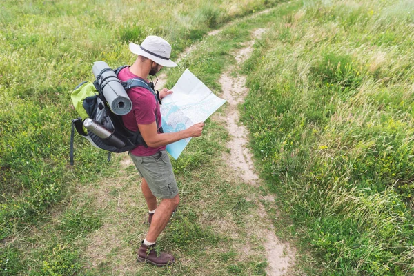 Voyageur masculin en chapeau avec sac à dos, thermos, tapis touristique en regardant la carte — Photo de stock