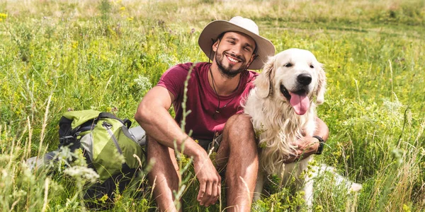 Viajero sentado con perro golden retriever en pradera verde - foto de stock
