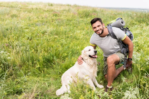 Viajante sorrindo com mochila e cachorro bonito sentado no prado verde — Fotografia de Stock