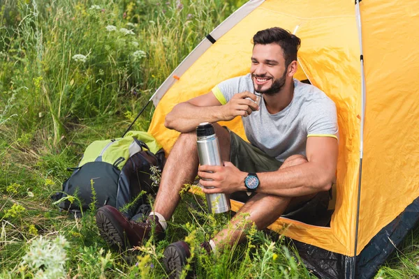 Voyageur souriant assis dans une tente jaune avec sac à dos et boire du café de thermos — Photo de stock