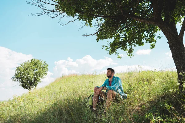 Junger Reisender mit Rucksack sitzt auf grüner Sommerwiese — Stockfoto