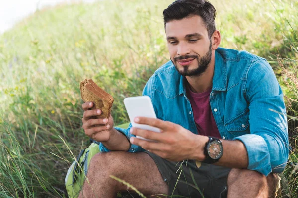 Traveler using smartphone and eating sandwich on summer meadow — Stock Photo