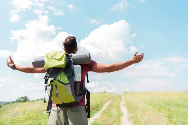Back view of hiker with backpack and tourist mat standing with outstretched hands on green meadow — Stock Photo