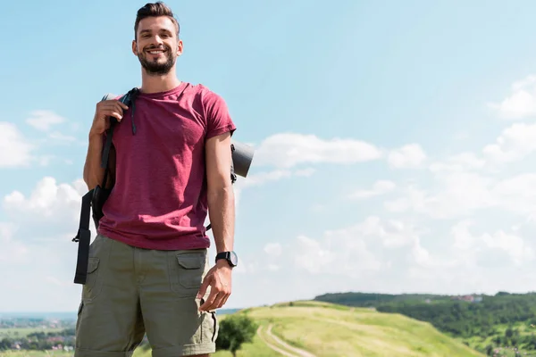 Lächelnder junger Wanderer mit Rucksack steht auf einer grünen Wiese mit blauem Himmel — Stockfoto