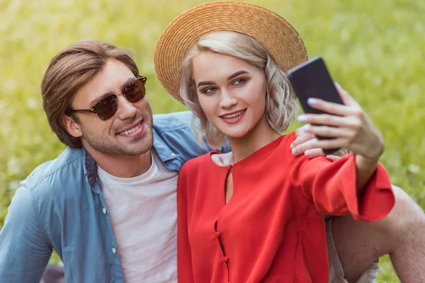 Sonriente pareja sentada en una manta en el parque y tomando selfie con smartphone - foto de stock