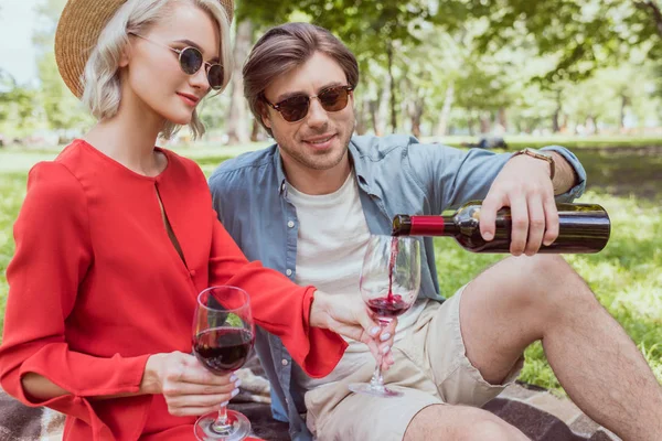Бойфренд наливает красное вино в бокалы из бутылки в парке — стоковое фото