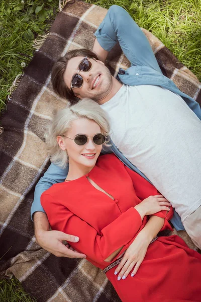 Visão de alto ângulo do casal em óculos de sol deitado e abraçando no cobertor no parque — Fotografia de Stock