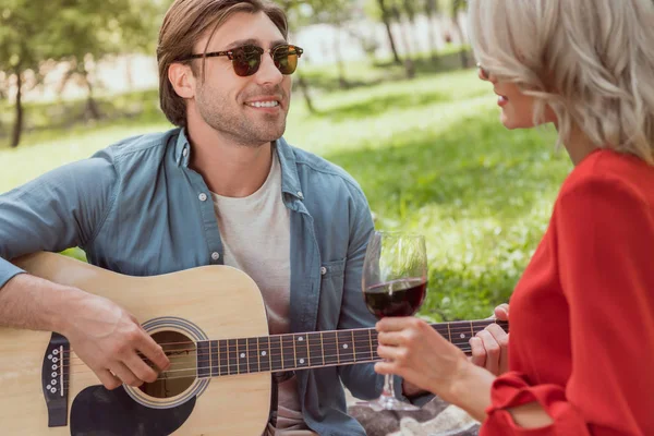 Guapo novio en gafas de sol tocando la guitarra acústica para novia en el picnic - foto de stock