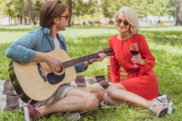 Красивый парень играет на акустической гитаре для девушки с вином на пикнике — стоковое фото