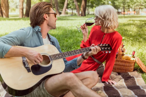 Guapo novio tocando la guitarra acústica para novia en el picnic en el parque - foto de stock