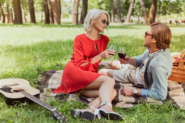 Feliz pareja acostada en la manta en el parque y tintineo con copas de vino tinto - foto de stock