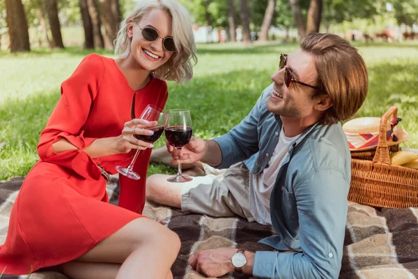 Happy couple lying on blanket in park and clinking with glasses of wine — Stock Photo