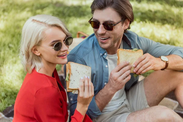 Couple in sunglasses eating sandwiches at picnic in park — Stock Photo