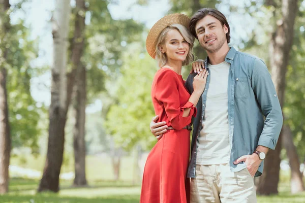 Novia sonriente y novio abrazándose en el parque y mirando a la cámara - foto de stock