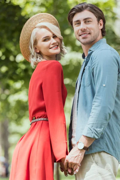 Vue latérale du couple tenant la main dans le parc et regardant la caméra — Photo de stock